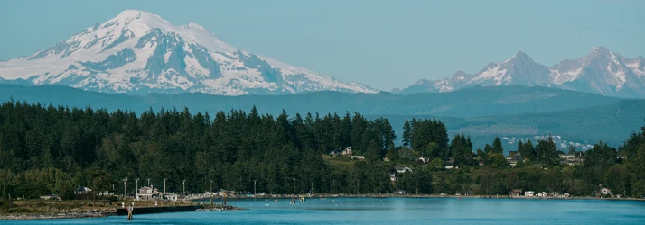 Mountain landscape with a snow-capped peak, forested shoreline, and calm water in the foreground.